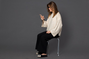 A girl in a light yellowish sweater and classic black trousers and glasses sitting on a chair, looks at the mobile phone in his hands