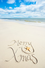 Merry Xmas message in handwritten calligraphic script in sand decorated with a natural starfish under sunny blue sky