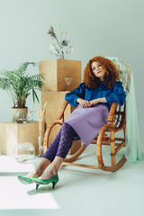 stylish redhead girl sitting in wicker chair near wooden boxes, glasses and dried flowers on grey
