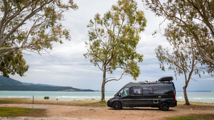 black camping car under trees at beach at Drepano Beach, Greece, with a green sea in the background
