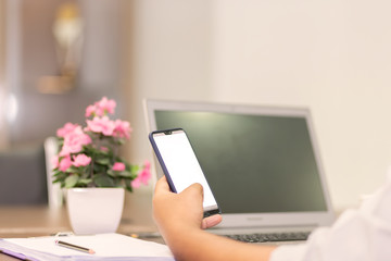 A man's hand is pressing on the smartphone screen on the table and the laptop is in the background. A business man eating coffee in a paper cup and pressing the phone with white screen.