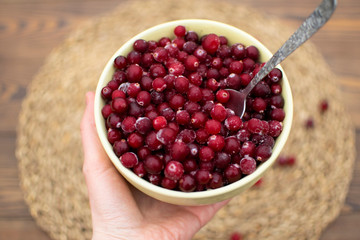 Female hand giving big bowl full of frozen cranberry with metal spoon inside at blurred wooden background with eco napkin, scattered berries, eco living