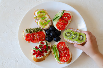 Children's hand with delicious fruit, vegetable sandwiches on a white round plate. Flat lay.