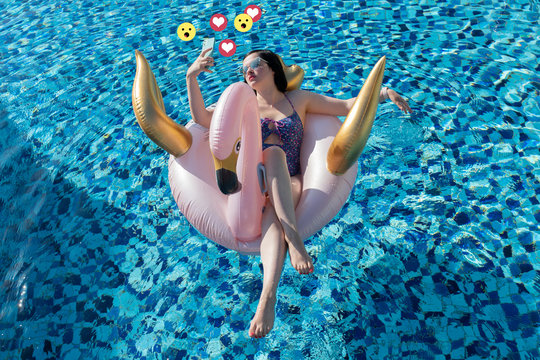 Social media influencer concept: young beautiful woman taking a selfie with mint smartphone on a swan / flamingo pool float in the blue swimming pool with heart and wow emoticons - Powered by Adobe