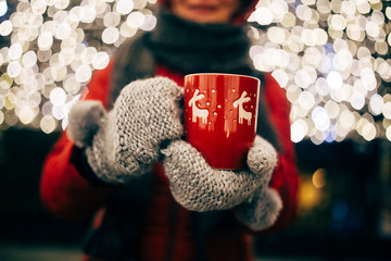 Christmas light and a woman holding in hand a red mug with hot drink - obrazy, fototapety, plakaty
