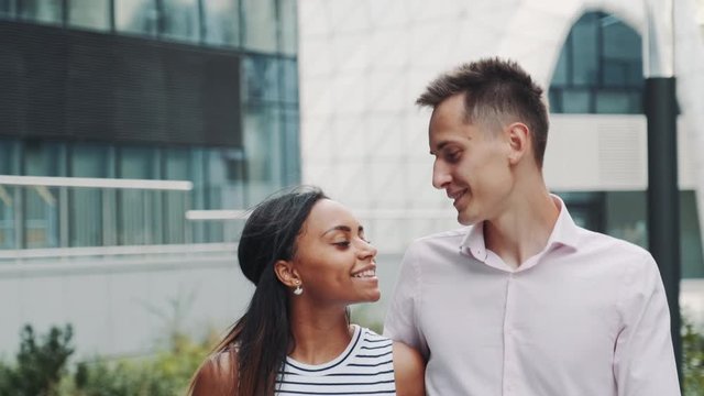 Close-up of cheerful multiracial couple in love kissing each other on date, speaking and hugging. They walking on the street with modern office building in the background.