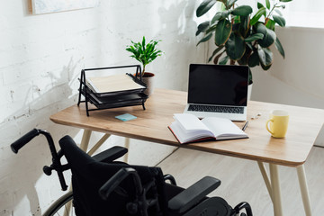 Workplace with open notebook, laptop and wheelchair by table