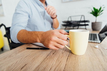 Cropped view of man in wheelchair drinking coffee at office desk