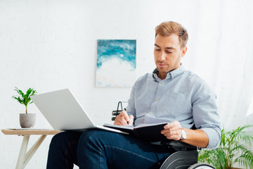 Man in wheelchair writing in notebook by desk with laptop
