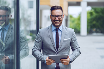 Handsome smiling caucasian businessman in gray suit and with eyeglasses holding tablet while standing outdoors and looking at camera. - Powered by Adobe