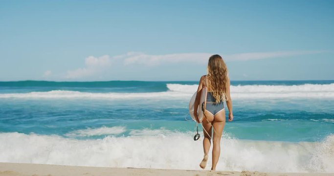 Attractive tan woman going surfing on a sunny day, walking out towards the ocean on the beach in slow motion, ocean spray beach day