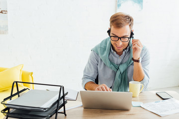 Smiling man in eyeglasses using headset and laptop at working table