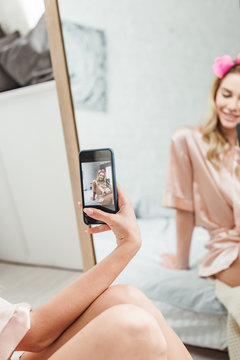 selective focus of happy woman with hair curlers holding smartphone while taking photo near mirror