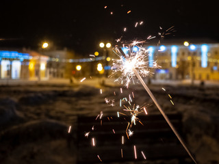 Sparkling sparkler on a city street in the dark in winter. In the background is urban festive...