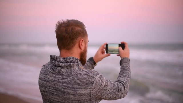 Handsome man with red beard in sweater on the beach take photo of sunset
