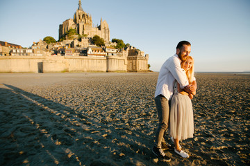 Young couple in love hugging in Europe on the beach next to Le Mont Saint-Michel tidal island in...