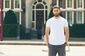 Fototapeta premium Hipster handsome male model with beard wearing white blank t-shirt with space for your logo or design in casual urban style
