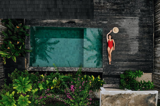 Aerial drone photo of happy Woman in red swimsuit relaxing near private pool with flowers and greenery around, Bali. Tropical background and travel concept.
