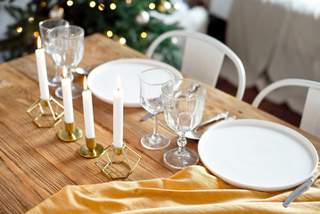 Table is served for a Christmas dinner in the living room, close-up view, table setting with a golden tablecloth, white plates, forks, knives, glasses for wine, candles in gold candlesticks, Christmas