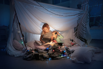 Fototapeta na wymiar Mother and daughter sitting in a teepee, reading stories with the flashlight in dark room with toys and pillows. Caucasian models. Home comfort, family, love, Christmas holidays, storytelling time.