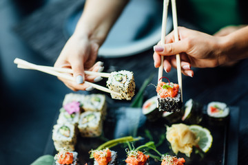Essstäbchen mit Sushi-Rollen mit Lachs und Frischkäse und Gurke auf schwarzem Teller. Nahaufnahme. Uramaki, Nori Maki oder Futomaki Sushi mit Forellenfilets, Sojasauce und Wasabi.