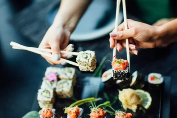 Foto auf Acrylglas Sushi-bar Essstäbchen mit Sushi-Rollen mit Lachs und Frischkäse und Gurke auf schwarzem Teller. Nahaufnahme. Uramaki, Nori Maki oder Futomaki Sushi mit Forellenfilets, Sojasauce und Wasabi.