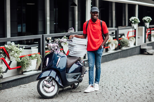 Busy african man, carries cardboard boxes with pizza, delivers to customers, poses on blue scooter, wears helmet, white t shirt and sportshoes, spreads legs, has happy expression.