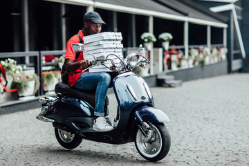 Young man courier poses on fast motorbike, carries rucksack, wears helmet, holding cardboard, looks aside with pleased expression. People, technology, transport.