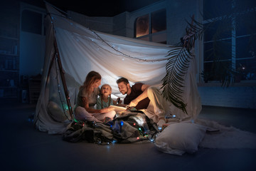Caucasian family sitting in a teepee, reading stories with the flashlight in dark room with toys...