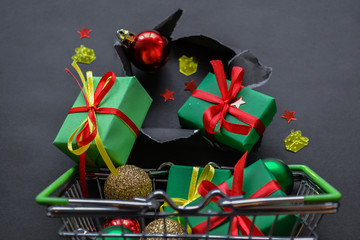 Gifts in green paper with red and yellow ribbons, confetti in the form of red stars and gold presents fall out a torn hole in the black paper into the metal basket full of presents and christmas balls