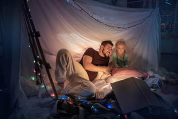 Obraz na płótnie Canvas Father and daughter sitting in a teepee, having fun, playing with the flashlight in dark room with toys and pillows. Look happy. Home comfort, family, love, Christmas holidays, storytelling time.