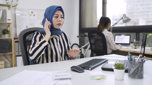 Busy serious puzzled muslim woman employee sitting in coworking shared office with diverse multicultural colleague holding mobile phone talking with client solving problem distantly. mad malay worker