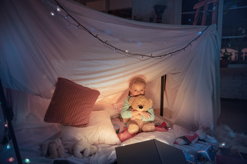 Little girl lying in a teepee, having fun, playing with the flashlight in dark room with toys and pillows. Look happy, holding teddy bear. Home comfort, childhood, Christmas holidays, storytelling. © master1305