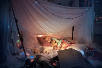 Little girl lying in a teepee, sleeping with the flashlight in dark room with toys and pillows. Look cute, holding teddy bear. Home comfort, childhood, Christmas holidays, sweet dreams time. © master1305