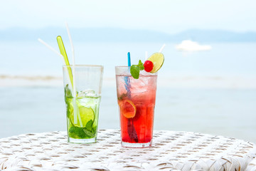 Two cocktails on the beach. Cold fancy colored summer drinks with lemon slices ,mint and cherry on a beach side table. Cold refreshment drinks with blurry beach scenery background.