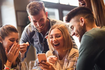 Fotobehang Groep jonge vrienden die plezier hebben in restaurant praten, lachen tijdens het dineren aan tafel en selfie maken. © Zoran Zeremski