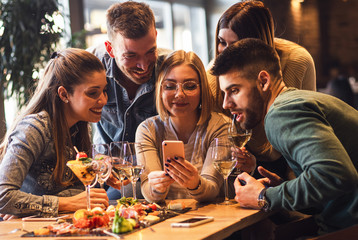 Group of young friends having fun in restaurant talking, laughing while dining at table and making selfie.	