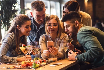 Fotobehang Group of young friends having fun in restaurant talking, laughing while dining at table and making selfie.  © Zoran Zeremski