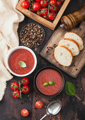 Ceramic bowl plates of creamy tomato soup with spoon, pepper and kitchen cloth on wooden background with box of raw tomatoes and bread.