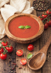 Wooden plate of creamy tomato soup with wooden spoon, pepper and kitchen cloth on wooden background with raw tomatoes.