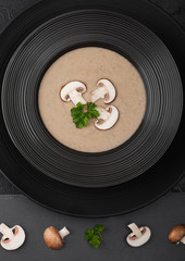 Black restaurant plate of creamy chestnut champignon mushroom soup on black table background with black stone board and fresh mushrooms.