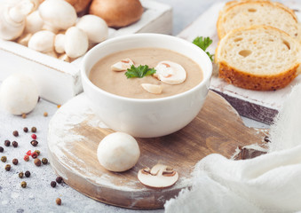 White bowl plate of creamy chestnut champignon mushroom soup on white kitchen background and box of raw mushrooms and fresh bread.