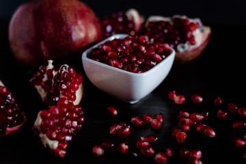 White bowl with pomegranate seeds and ripe pomegranate on dark wooden background.