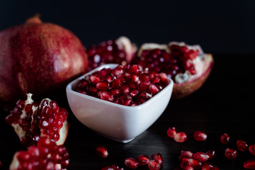 White bowl with pomegranate seeds and ripe pomegranate on dark wooden background.