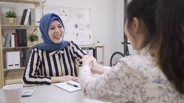 charming smiling young muslim female insurance agent in hijab shaking hands with lady client while meeting in office. malay woman investment adviser handshaking with girl customer after great deal.
