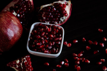 White bowl with pomegranate seeds and ripe pomegranate on dark wooden background.