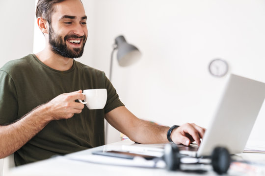Image of caucasian smiling man typing on laptop and drinking coffee