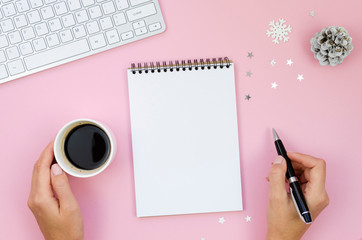 Female Workplace with Keyboard Laptop Computer, Coffee cup, and Christmas Decoration. Woman's hand holding pen and writing letter. Merry Christmas mail concept mockup with blank spiral notepad