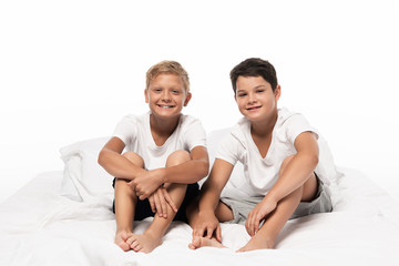 Fototapeta na wymiar two cheerful brothers sitting on bed and looking at camera isolated on white
