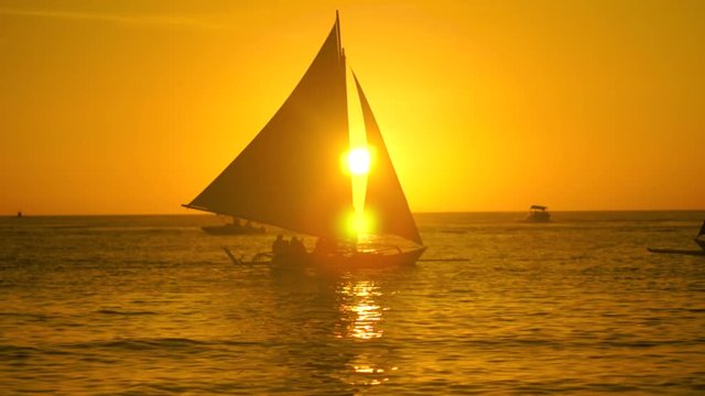 The silhouette of the boat with people sailing along the horizon. The boat sails from left to right and crosses the sun in the middle of the frame. Bathed in the red rays of the setting sun
